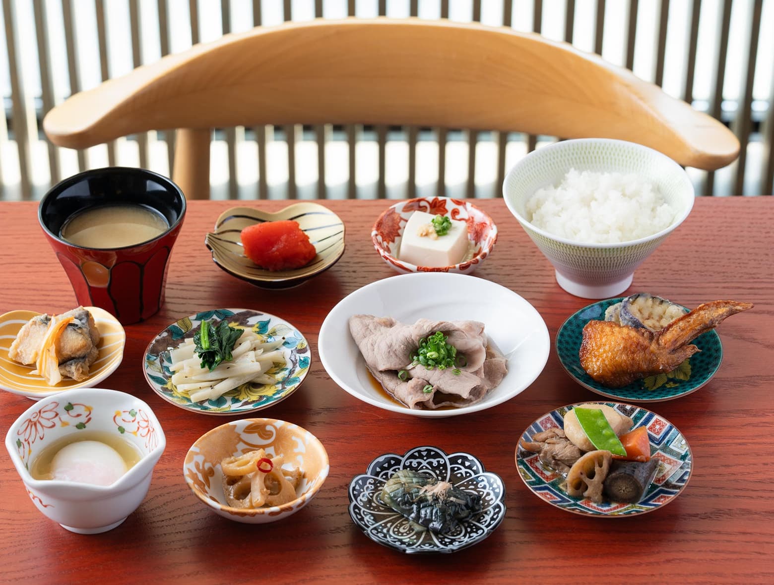 A variety of small, colorful Japanese dishes are neatly arranged on a wooden table, featuring rice, miso soup, and various savory items.
