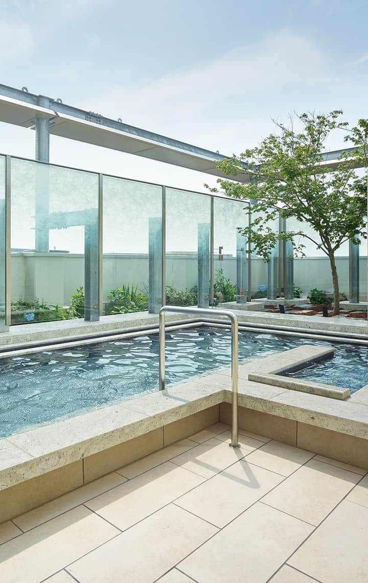An outdoor rooftop large public bath with clear water, surrounded by glass panels, small trees, and stone accents.