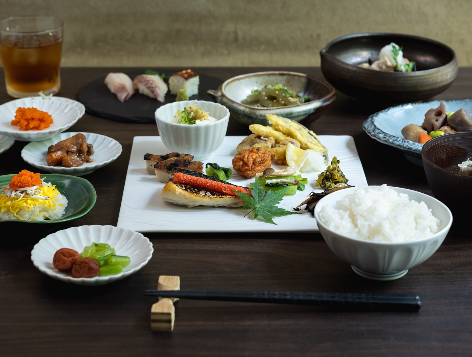 木桌上摆放着菜色丰富的日式早餐，包括烤鱼、天妇罗、米饭、寿司、小配菜和传统腌梅。