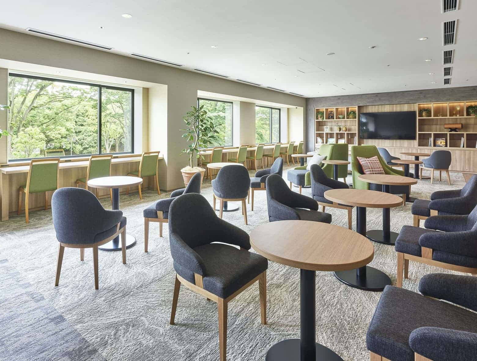 편안한 회색 의자, 원형 테이블, TV, 그리고 푸른 녹음을 감상할 수 있는 카운터 좌석이 마련된 밝고 모던한 분위기의 호텔 라운지.