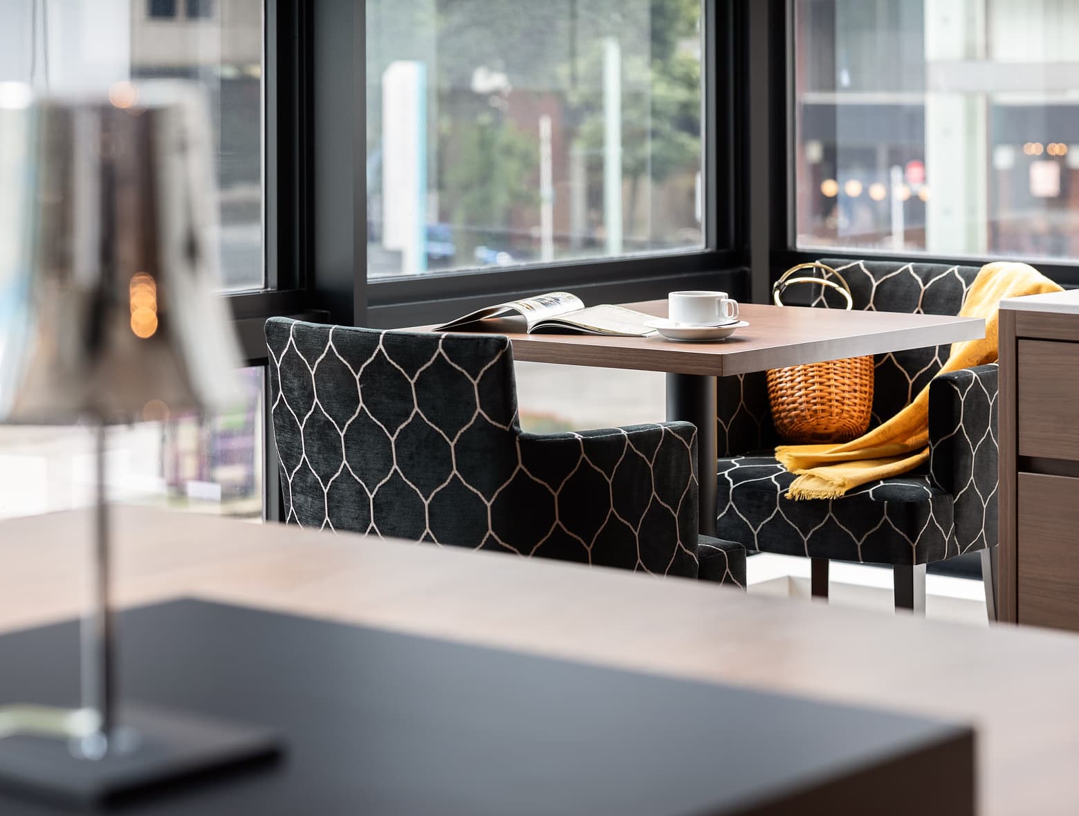 검정색과 금색 기하학적 패턴의 의자, 커피잔이 놓인 작은 테이블, 노란색 담요가 있는 창문 옆 아늑한 라운지 좌석 공간.
