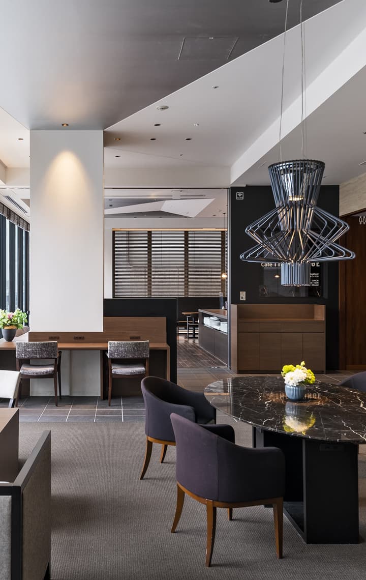 도시의 거리가 보이는 대형 창문, 어두운 컬러 톤의 우드 가구, 대리석 테이블, 현대적인 조명을 갖추고 있는 모던한 분위기의 로비.