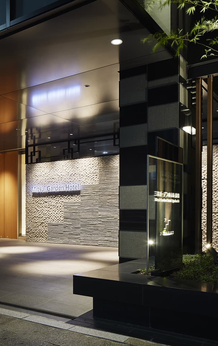 밤이 되면 조명이 켜진 대나무 벽이 돋보이는, 어두운 컬러 톤의 석재와 유리로 마감된 미쓰이 가든 호텔 교바시의 모던한 외관 입구.