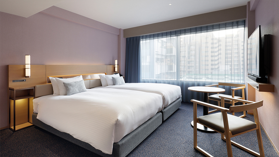 The Deluxe , 2 beds room features two beds, a small round table with chairs, a wall-mounted TV, and ample natural light from a large window.