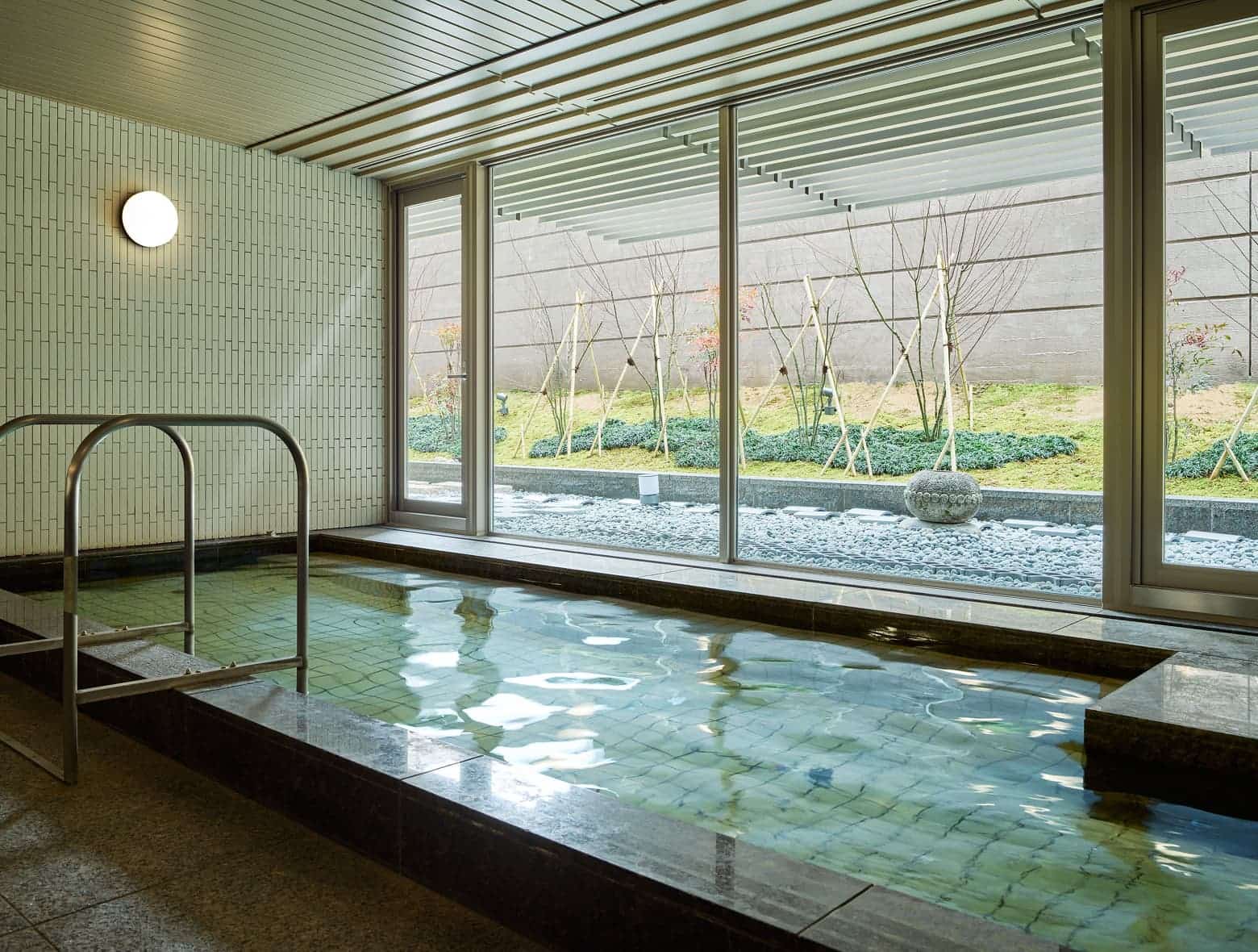 밝은 색 타일 벽, 테두리를 어두운 톤의 석재로 마감한 욕탕, 자갈과 잔디가 깔린 야외 공간이 보이는 대형 통창이 있는 세련되고 현대적인 실내 대욕장.