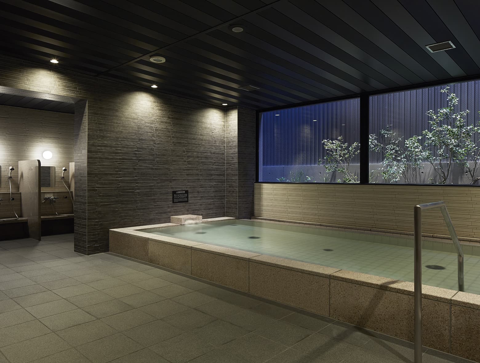 차분한 색감의 벽과 바닥 타일, 테두리가 석조로 된 욕탕, 샤워 공간, 식물이 있는 야외 정원이 보이는 긴 창이 특징인 대욕장.
