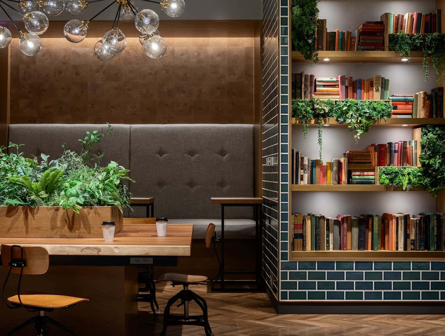 现代而温馨的酒店贵宾休息室配有共享桌子、嵌入式卡座以及摆满书籍和茂盛植物的书架。