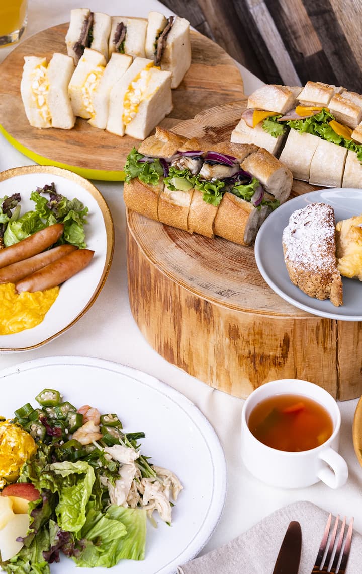 早餐特写，包括三明治、糕点、香肠炒鸡蛋和各种饮料，盛放在素雅的木质托盘和白色餐盘中。