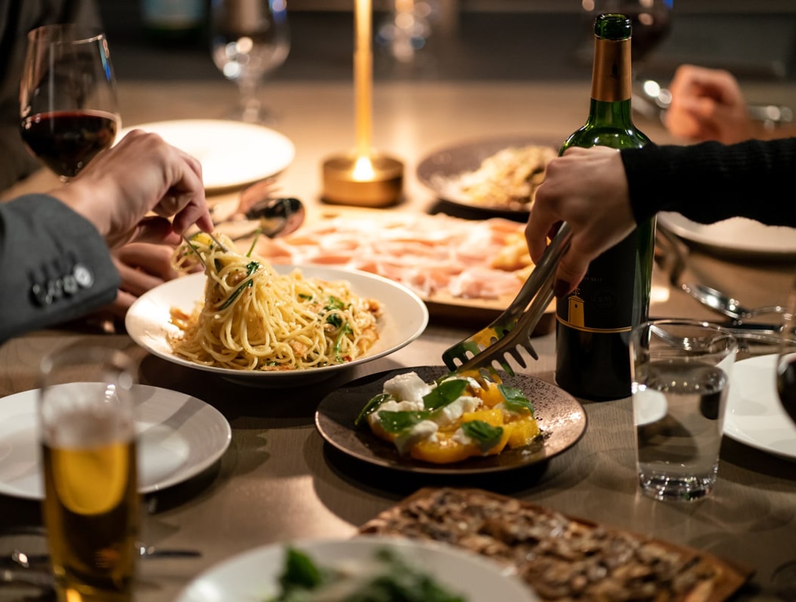Pasta, fruit, and deli meats are arranged on plates next to wine bottles well-lighted by candles, showcasing the dinner menu at BALCÓN TOKYO.