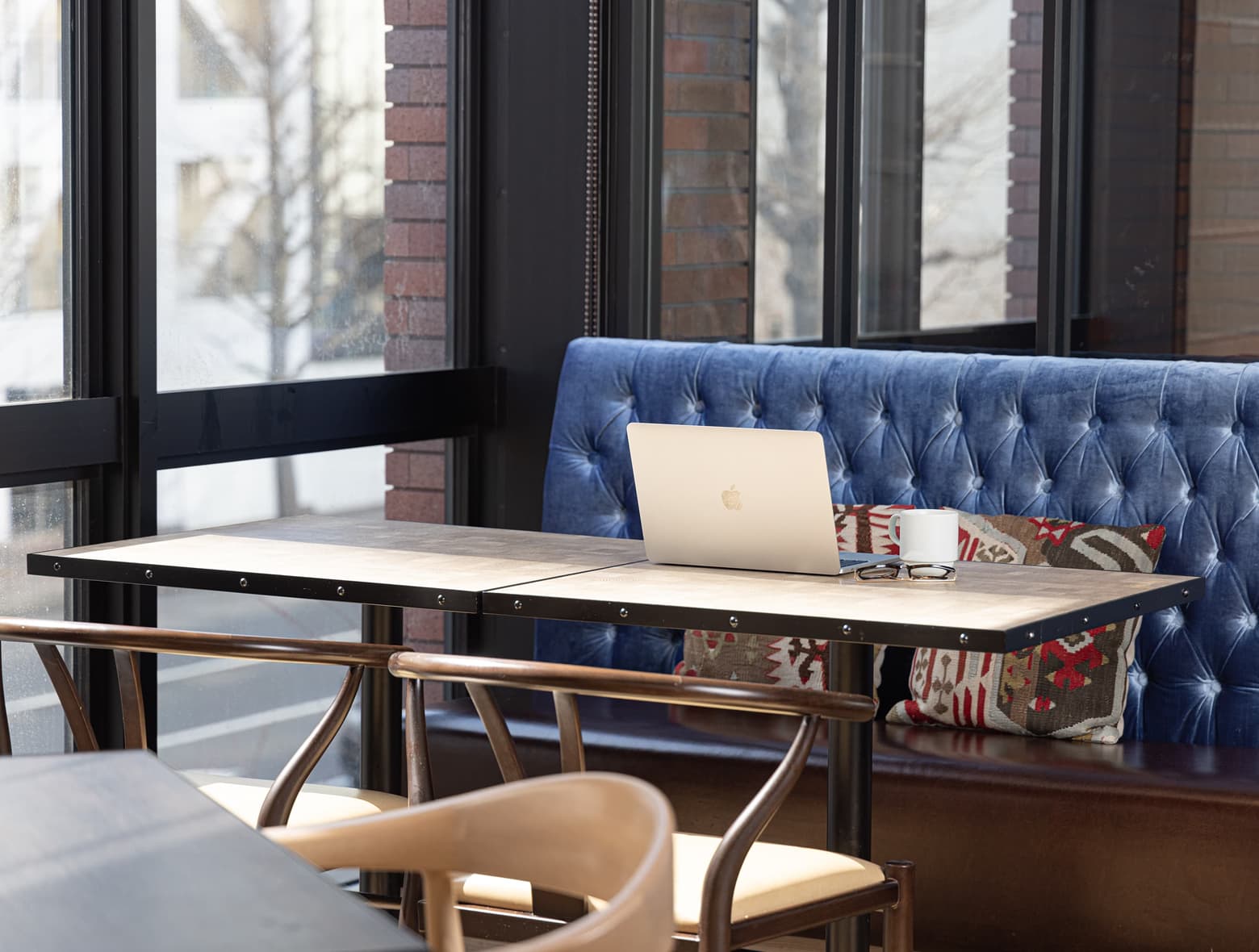 목재 테이블 위에 노트북이 놓여 있고, 파란색 벨벳 벤치 의자와 무늬 있는 쿠션, 커다란 창이 있는 호텔 라운지.