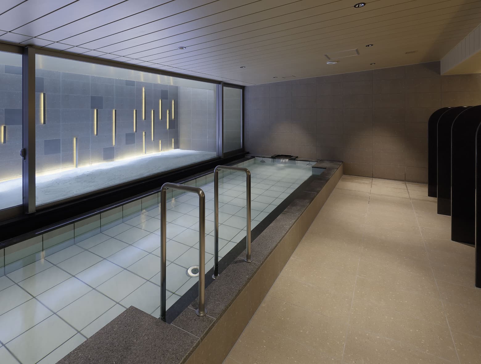 타일 욕탕에서 수직 조명과 석재 질감이 어우러진 장식 벽을 감상할 수 있는 현대적인 대욕장.