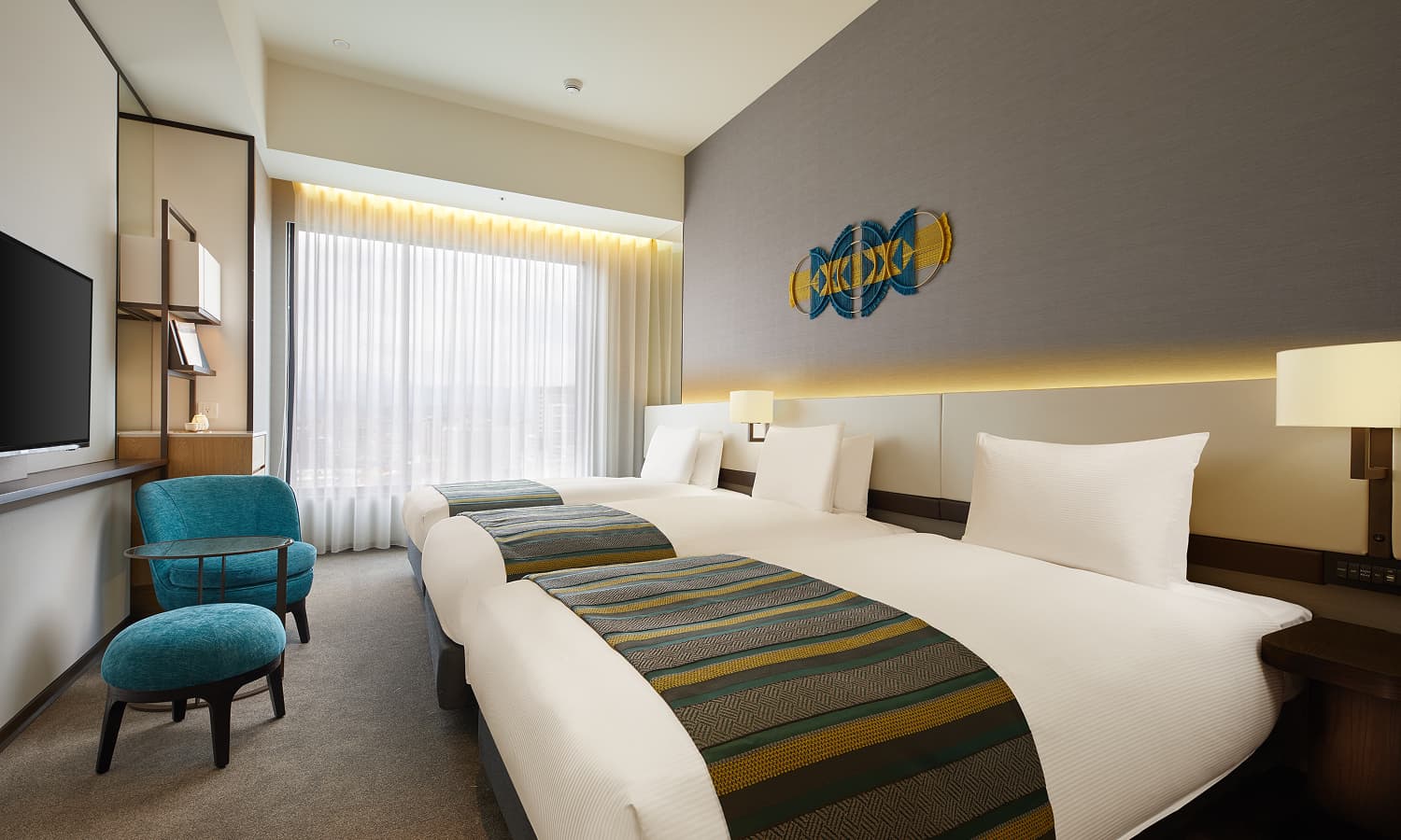 디럭스 트리플룸에는 침대 3개, 침대 헤드보드 위에 파란색과 노란색의 기하학적 무늬 직조 장식, 청록색 안락의자, 큰 창이 있습니다.