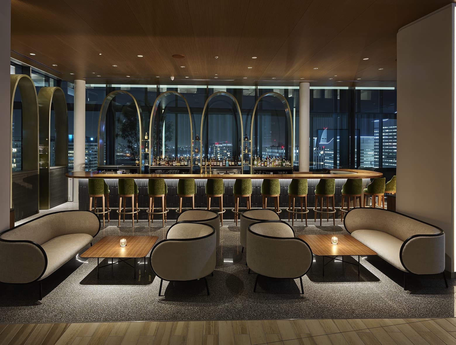 녹색 벨벳 의자와 곡선형 좌석, 그리고 도시의 스카이라인이 내려다보이는 긴 카운터가 있는 세련된 고급 호텔 바의 저녁 모습.