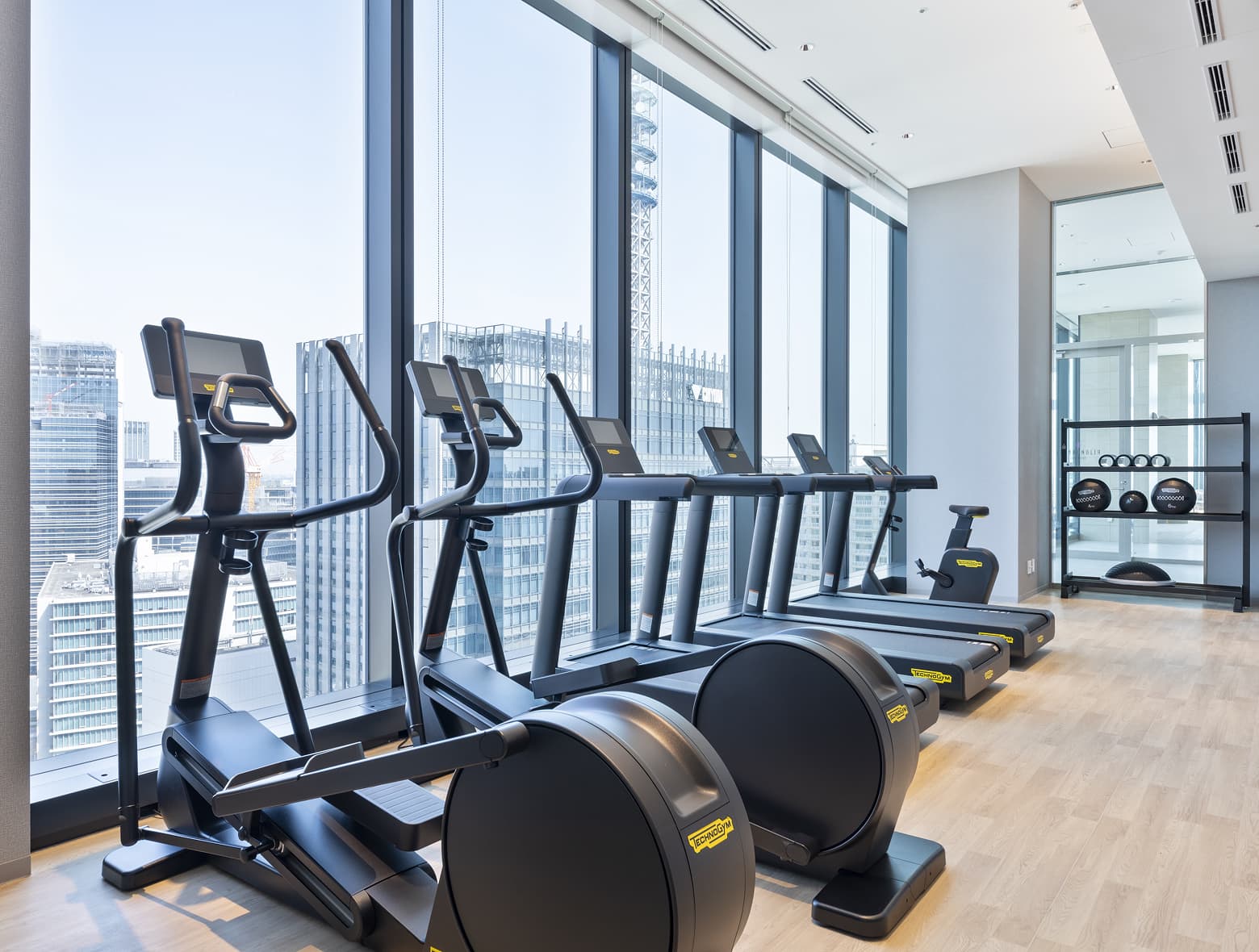 런닝머신과 일립티컬 등 유산소 운동기구가 완비된 현대적인 피트니스 룸으로, 모든 기구가 탁 트인 도시 전망을 감상할 수 있는 큰 창을 마주보고 있습니다.