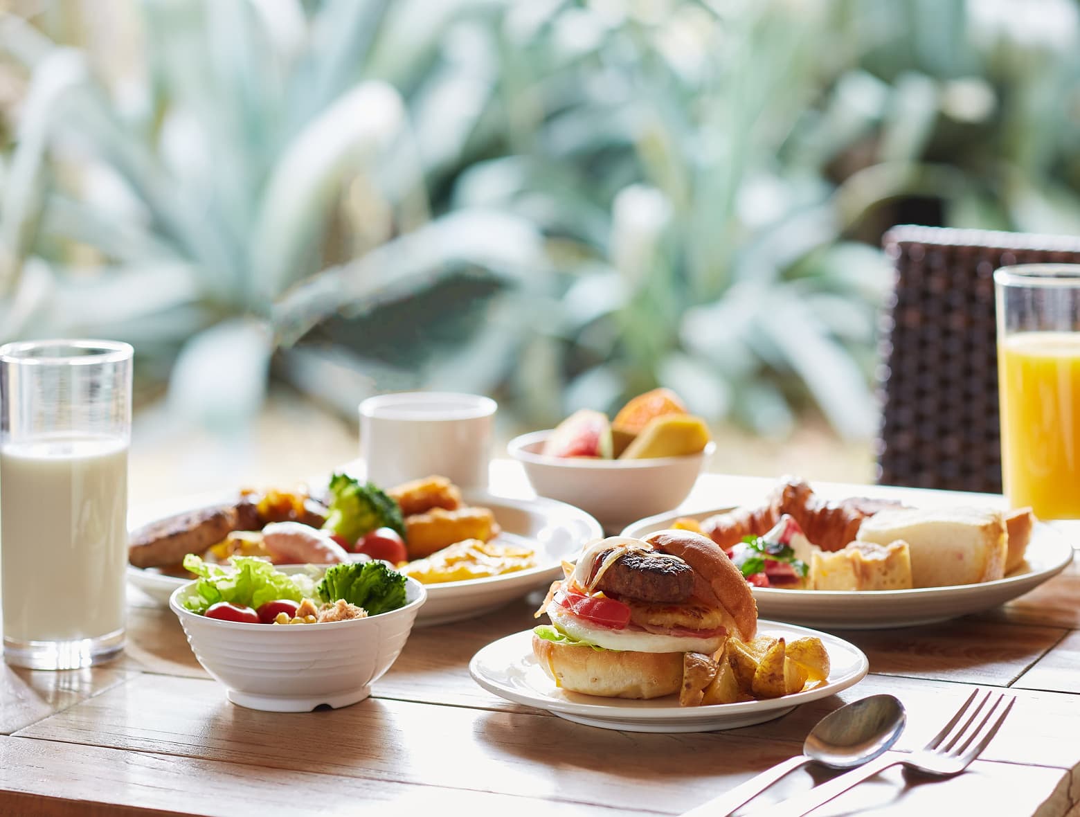 A breakfast spread on a wooden table, featuring a small sandwich, salad, sausage, and glasses of milk and orange juice.