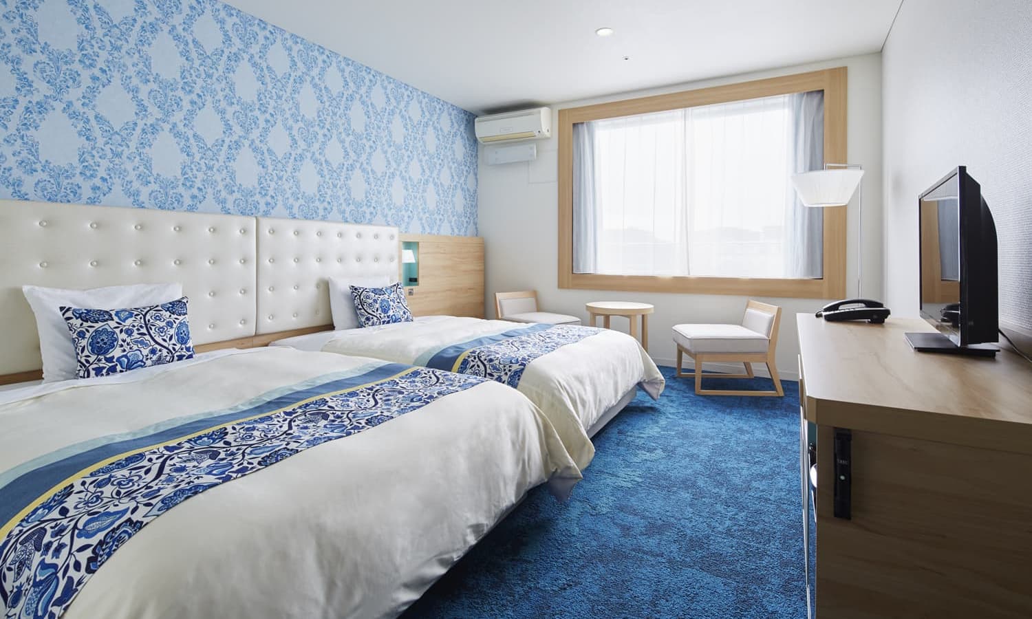 The Superior, 2 beds room features two beds, a blue and white patterned accent wall, a vibrant blue patterned carpet, and a large window for natural lighting.