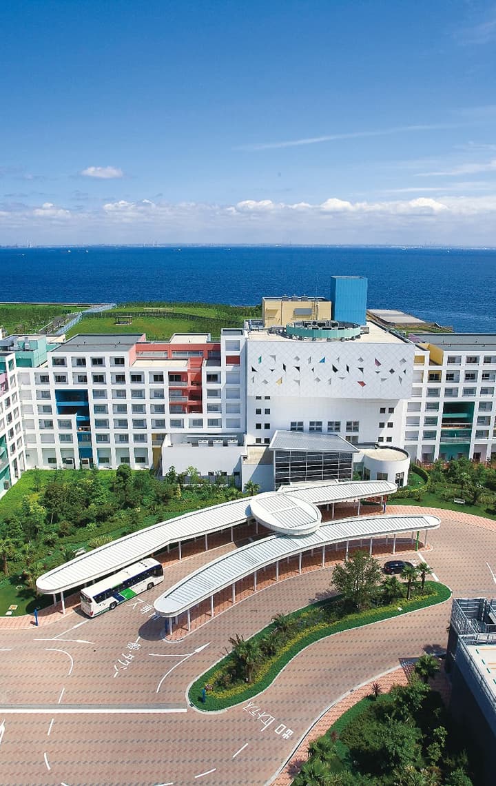A large, colorful seaside resort hotel with a white facade and curved entrance awning, surrounded by green landscape and the deep blue ocean.