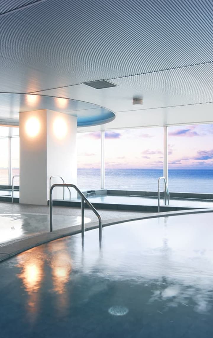 A large indoor public bath with dark stone edges and a panoramic window overlooking the vast, blue ocean and horizon.